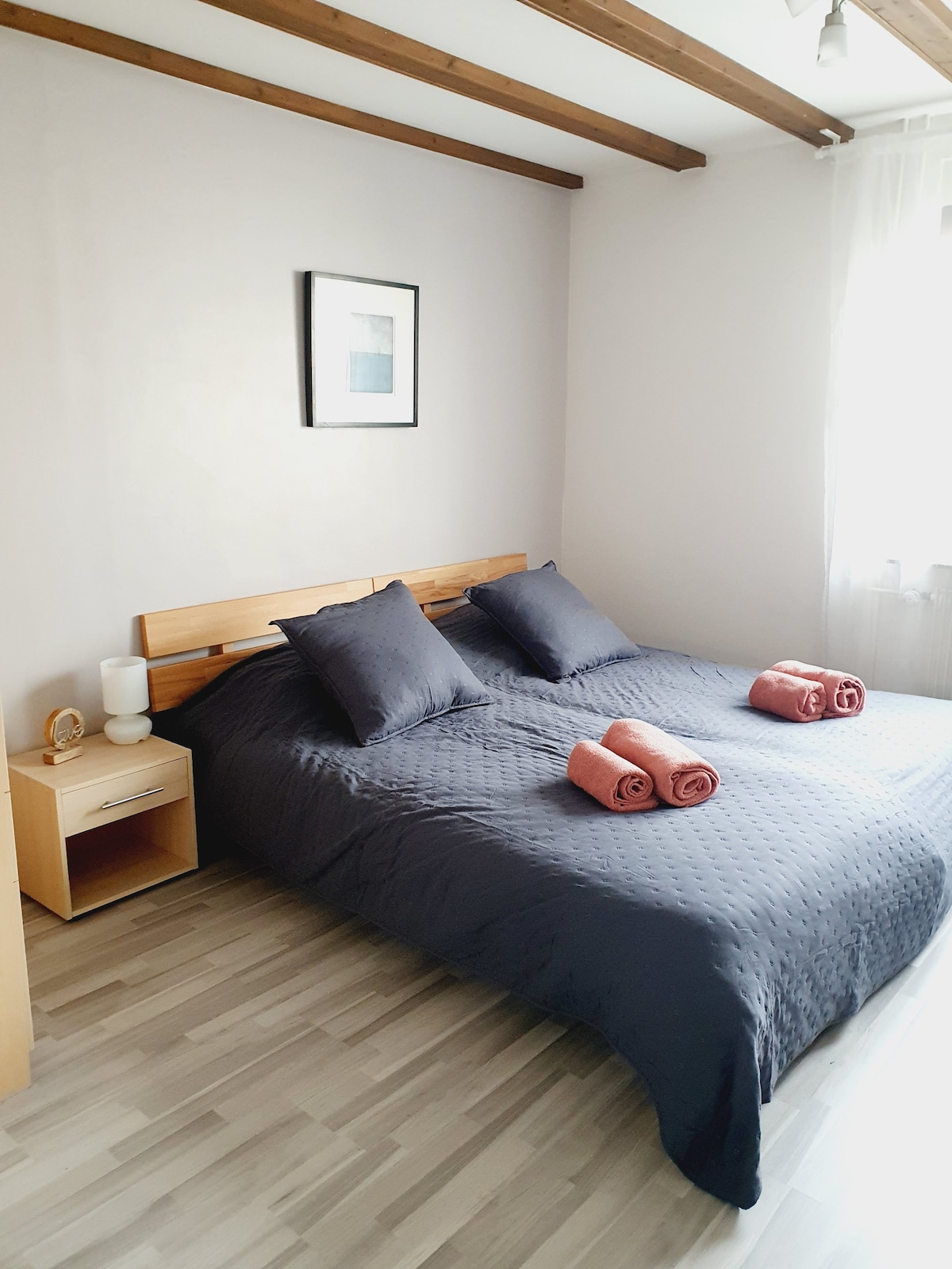A spacious bedroom features a large bed with dark bedding and two accent pillows. Two rolled towels are placed at the foot of the bed. A bedside table holds a lamp and a decorative piece. Natural light enters through a window adorned with sheer curtains.