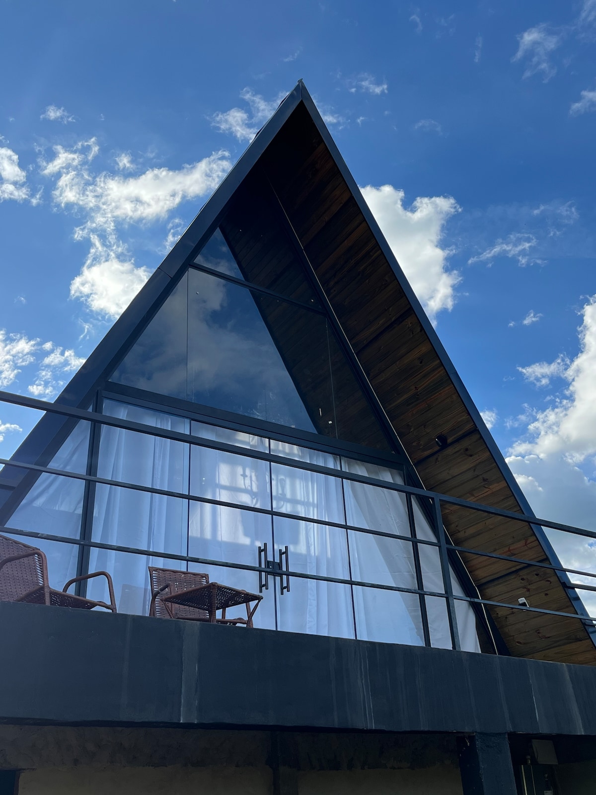 The exterior of the cabin features a striking A-frame design, highlighted by large glass panels that reflect the sky. A wooden overhang complements the sleek lines, while two rattan chairs sit on the balcony, inviting relaxation with a view.
