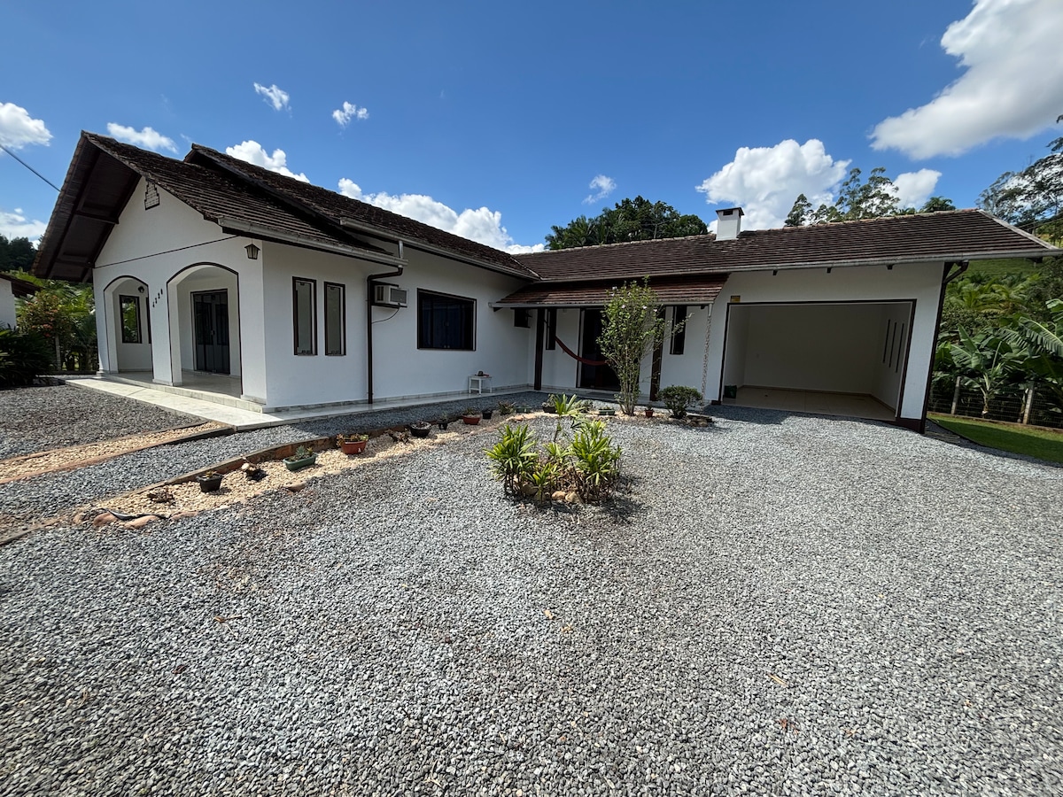 The exterior of a spacious house is showcased, featuring a gravel driveway and manicured gardens with native plants. The structure is surrounded by greenery, including trees and shrubs, and includes a covered garage. A clear blue sky and white clouds provide a bright backdrop.