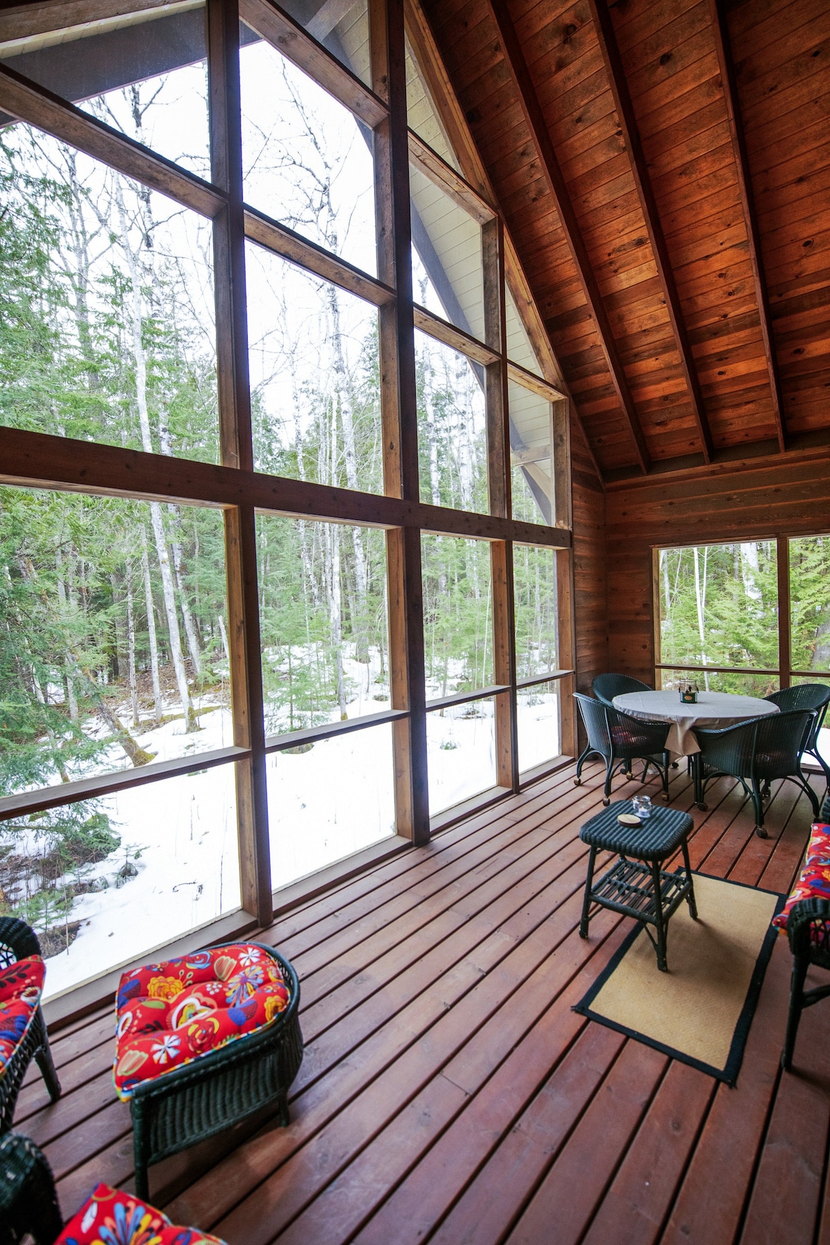 A spacious screened porch with large windows showcases natural surroundings, allowing for views of the forest and snow-covered ground. Comfortable seating with colorful cushions is arranged around a small table, creating a welcoming space for relaxation or dining.