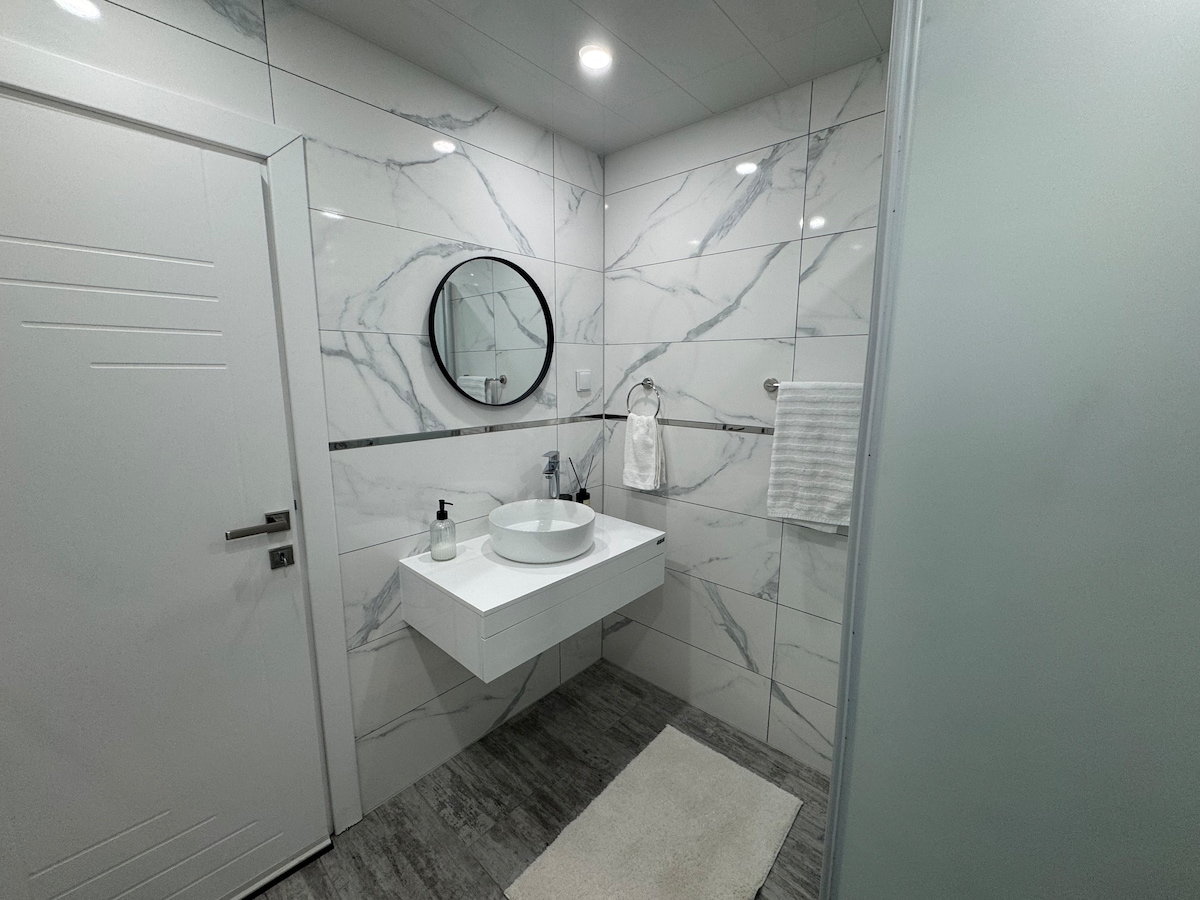 A modern bathroom features sleek white and grey marbled tiles and a minimalist floating vanity. A round mirror hangs above a white sink, which is accompanied by neatly arranged toiletries. Soft lighting enhances the space, and a fluffy rug is positioned on the floor.