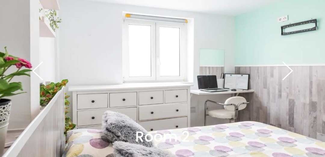 A bright room features light-colored walls and wooden accents. A spacious bed with a patterned blanket is visible, complemented by a white dresser and a desk with a chair. Natural light enters through a window, enhancing the room's airy feel.