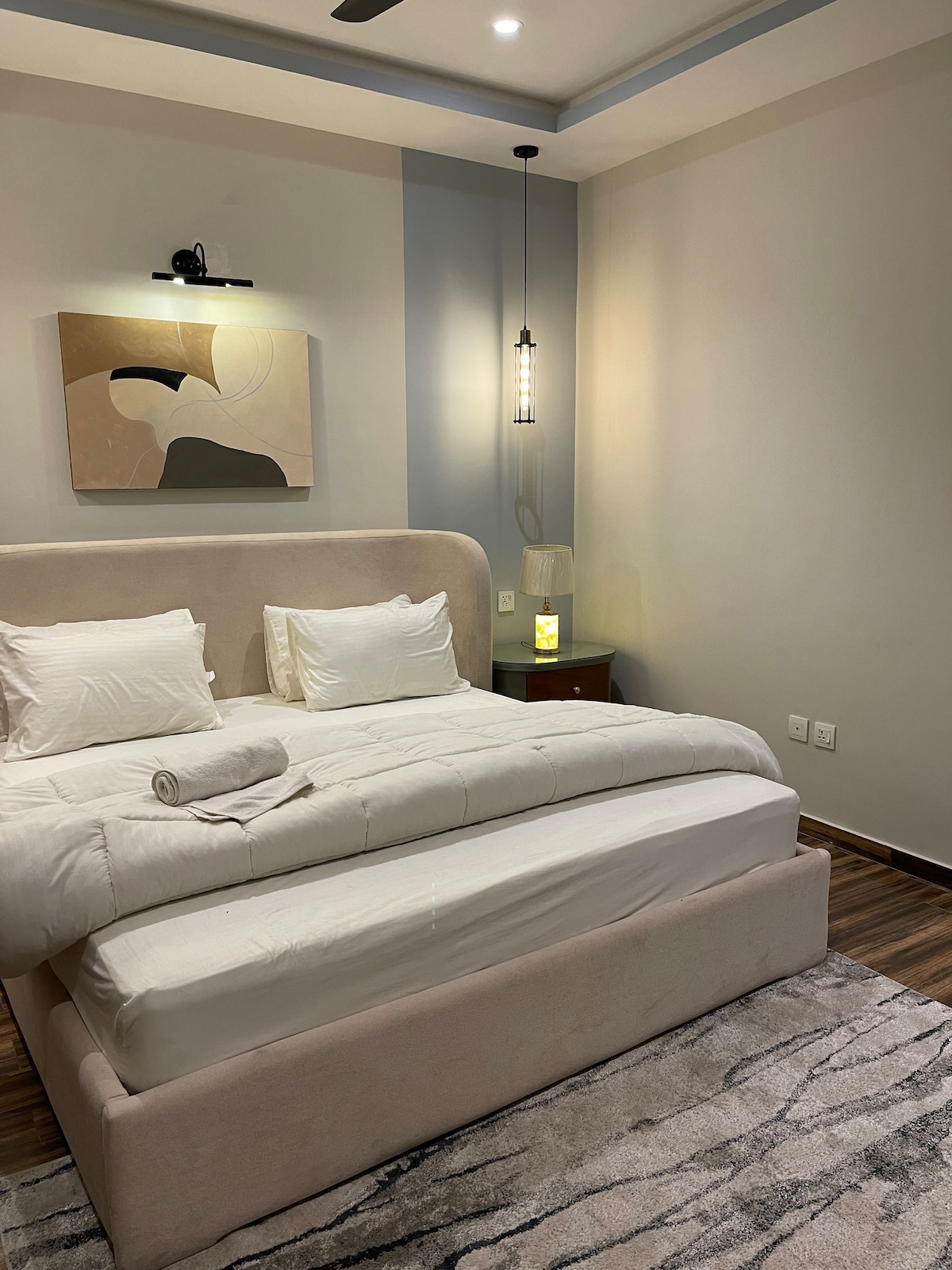 A serene bedroom features a plush bed with soft white linens and decorative pillows. A contemporary artwork piece is mounted above the bed, while a bedside table holds a softly glowing lamp. Warm light enhances the neutral color scheme and wood flooring.