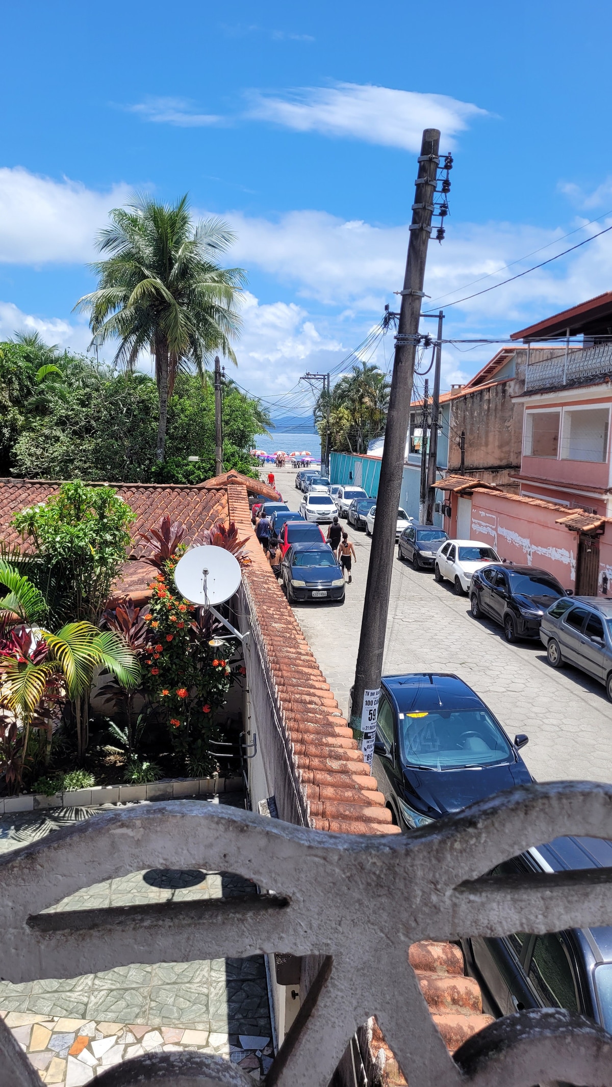A view from a balcony captures a serene street scene, lined with palm trees and various vehicles parked on both sides. In the distance, the beach is visible, framed by clear blue skies and fluffy white clouds, suggesting a pleasant atmosphere.