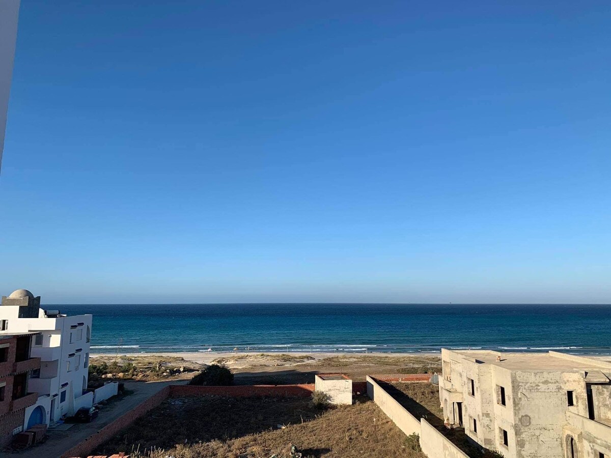 A panoramic view of the beach and the sea is captured, showcasing the expansive horizon with clear blue skies. The shoreline is framed by low-rise buildings, providing a sense of proximity to the water. The serene landscape creates a peaceful atmosphere.