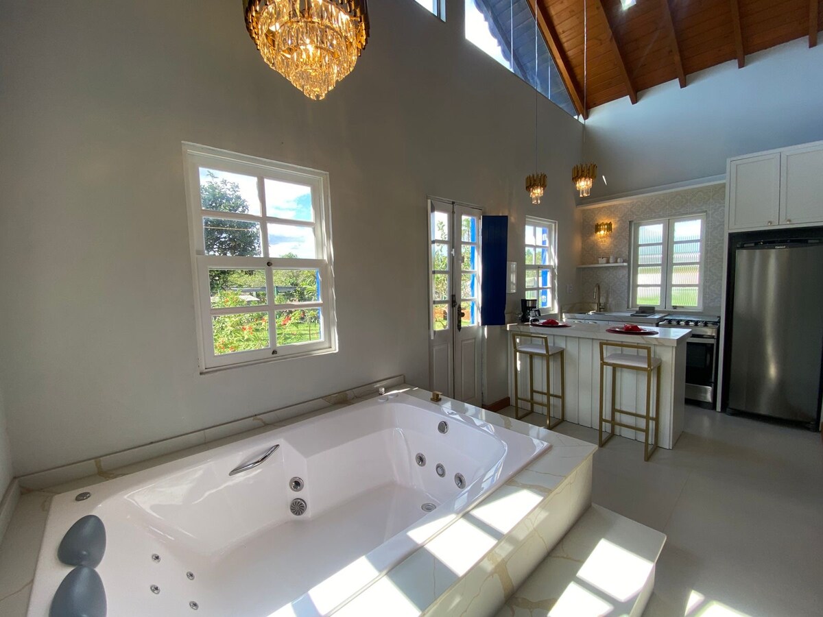 A bright and airy space features a large jacuzzi in the foreground, complemented by a modern kitchen area with bar stools. Tall windows allow for natural light to fill the room, highlighting the clean lines and light-colored decor.