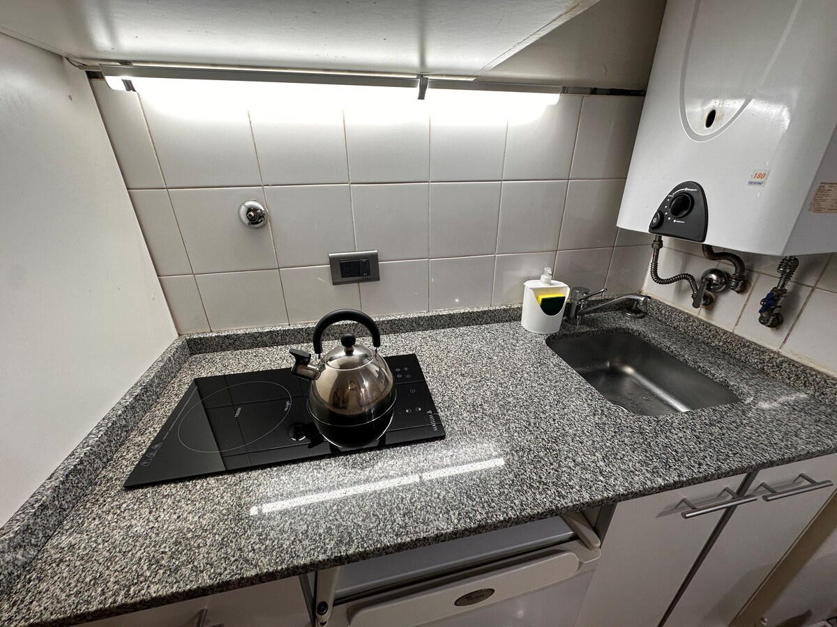 A compact kitchen space is presented with a grey granite countertop. An electric kettle sits on a black induction cooktop, alongside a small sink. Soft lighting illuminates the white tiled walls, while modern appliances are discreetly integrated within the cabinetry.