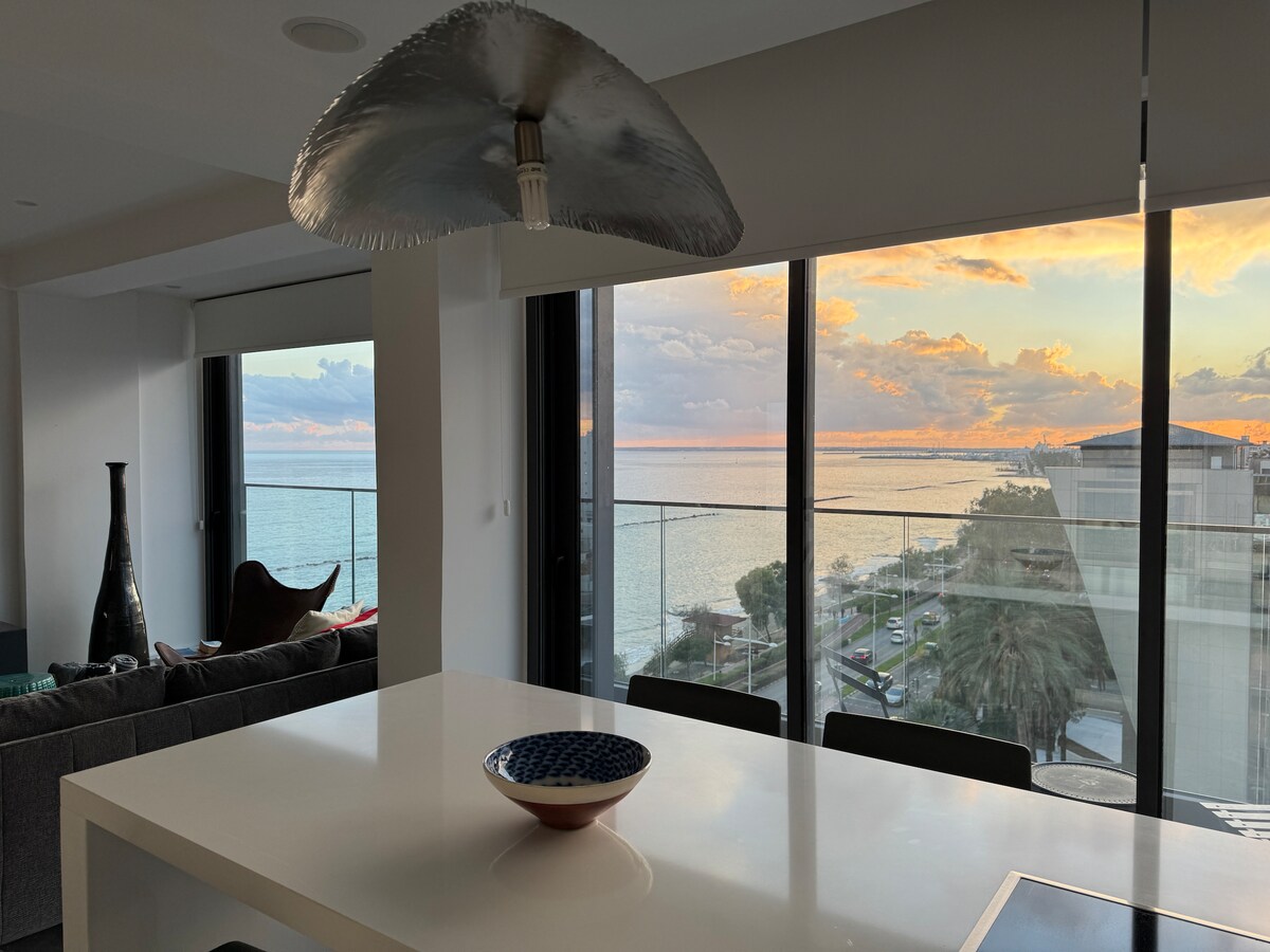 An open-concept living area is highlighted by large floor-to-ceiling windows showcasing sweeping ocean views. A sleek dining table is centered under an elegant pendant light, with a decorative bowl positioned invitingly at its center. Soft furnishings add to the modern atmosphere of the space.