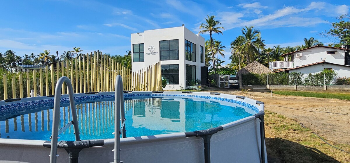 A round above-ground pool is surrounded by a grassy area, with a modern white building visible in the background. The structure features large glass windows and is framed by palm trees against a clear blue sky.