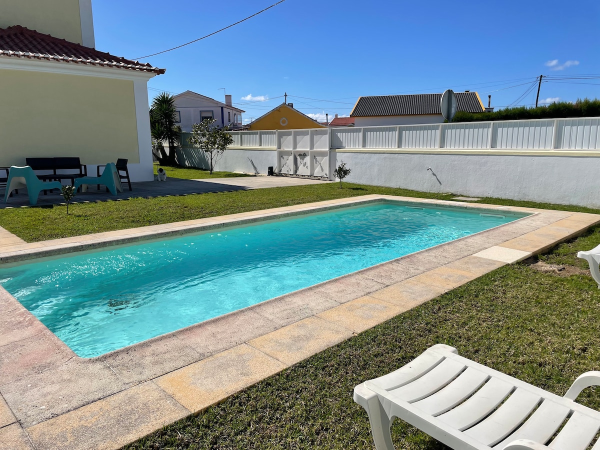 A private pool is set within a manicured green lawn, bordered by a light-colored stone patio. Sun loungers are positioned around the pool, inviting relaxation. The clear blue sky and distant houses provide a serene backdrop to this outdoor space.