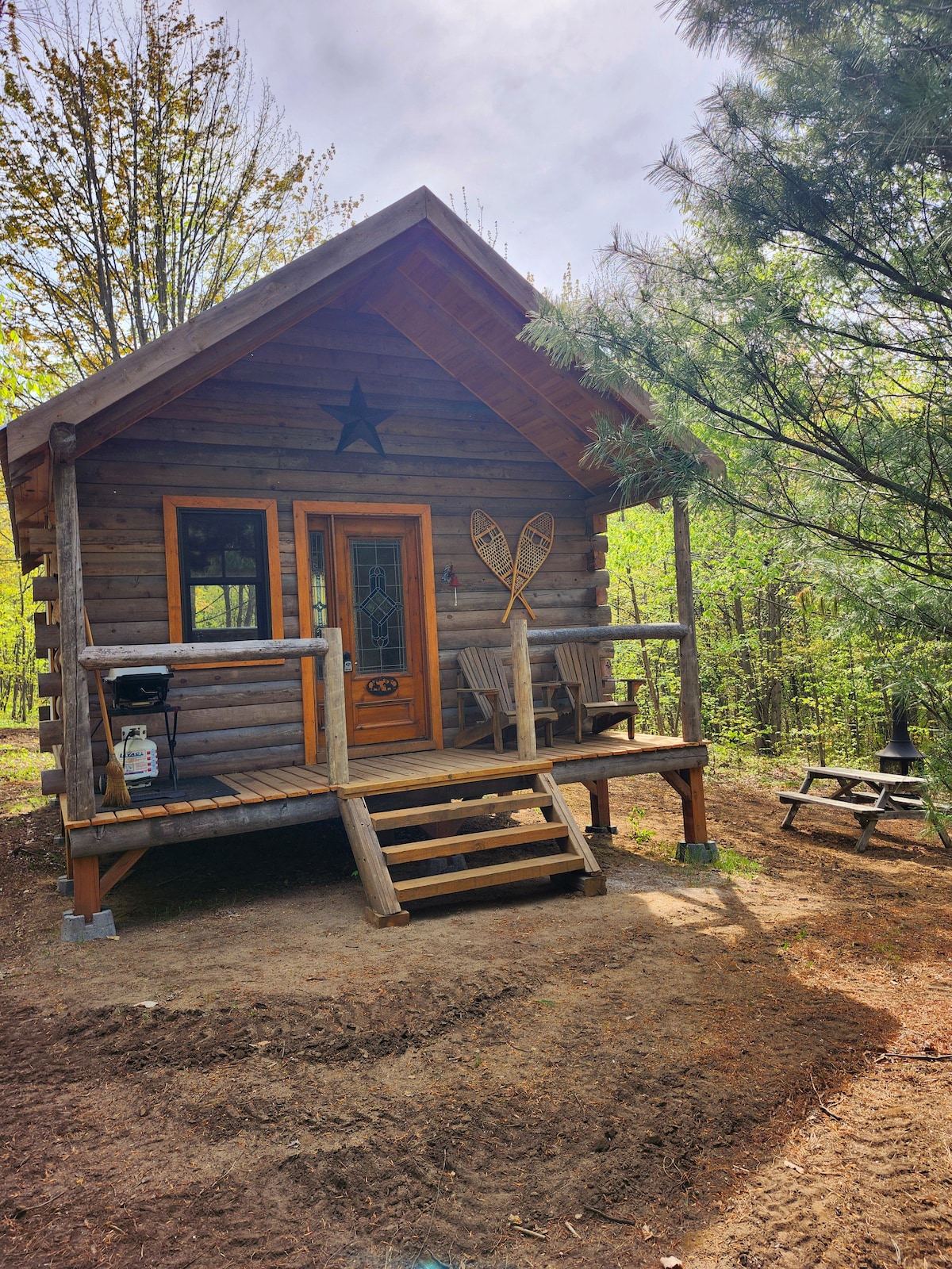 A rustic cabin crafted from logs is surrounded by trees, featuring a welcoming porch with two wooden chairs and a small table. A set of snowshoes decorates the wall beside the entrance, and steps lead up to the front door.