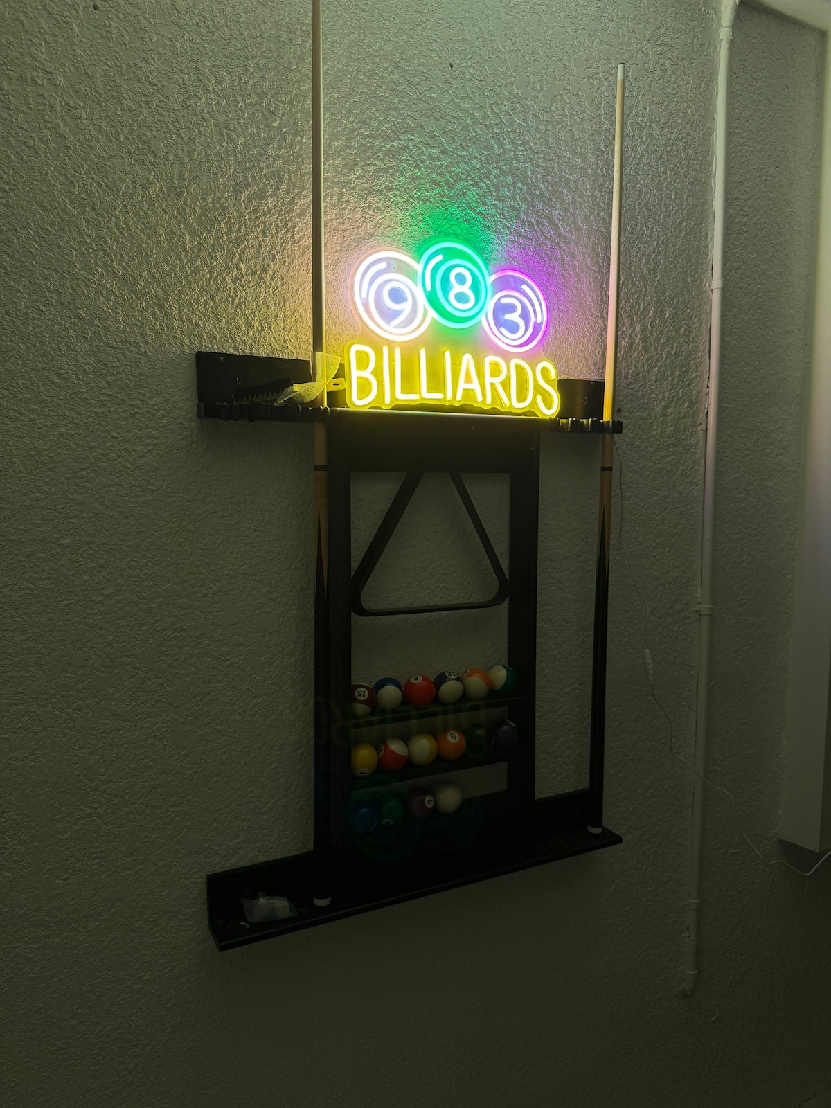 A neon sign displaying the word 'Billiards' is mounted on the wall, featuring vibrant lights in green, blue, and red. Below, a rack holds a full set of colorful billiard balls, neatly arranged in a triangular formation.
