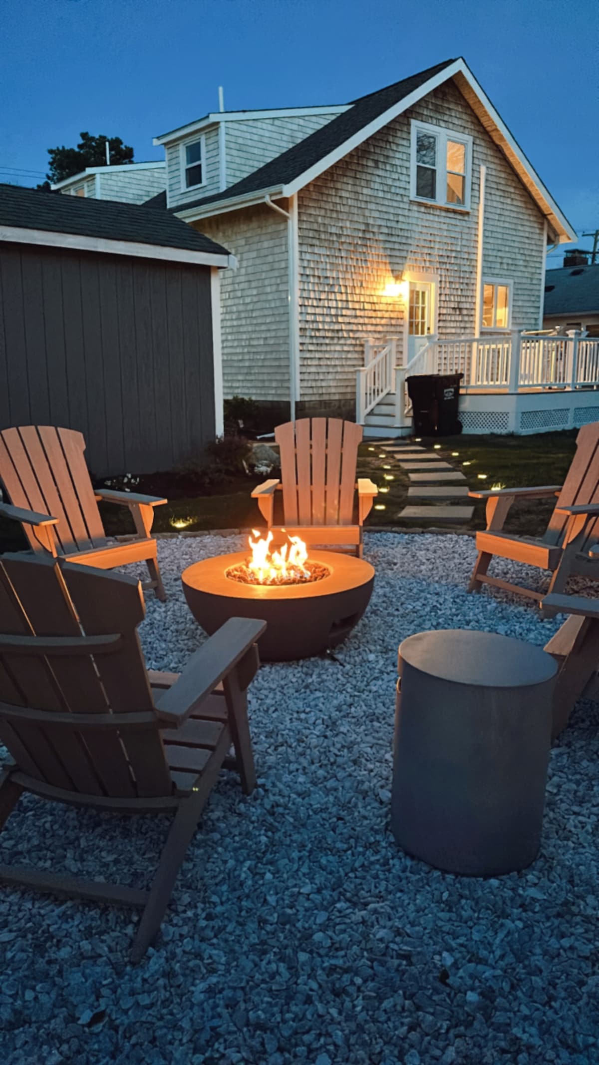 An inviting outdoor space features five adirondack chairs arranged around a circular fire pit, glowing warmly. Soft lights illuminate the exterior of the cottage in the background, highlighting the textured shingle siding and porch railing, with a gravel surface underfoot.