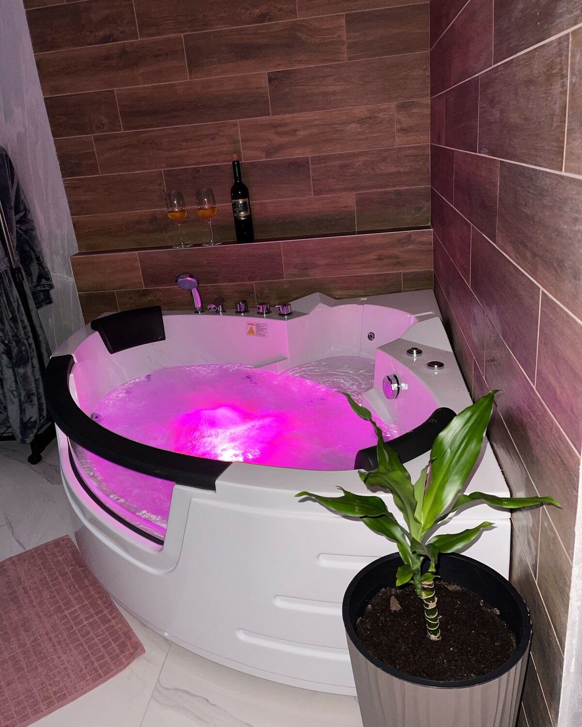 A circular hot tub is set against a backdrop of wooden wall panels. The tub features ambient lighting that creates a relaxing atmosphere. To the side, a small plant is placed in a modern pot, complementing the soothing environment. Two glasses of champagne sit on the shelf nearby.