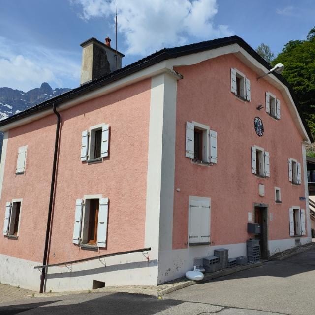 The exterior of a two-story building is shown, characterized by a soft pink façade and white shutters. A clock is visible near the roof, and the structure is framed by greenery and distant mountains. A sloping pathway leads to the entrance, providing accessibility.