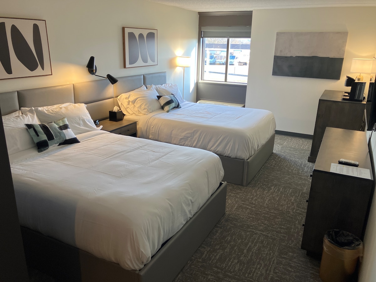 The room features two spacious queen beds with crisp white linens. A large window allows natural light to fill the space, while bedside lamps provide additional lighting. A functional coffee maker is positioned on a dark wood dresser, enhancing the room's practicality.