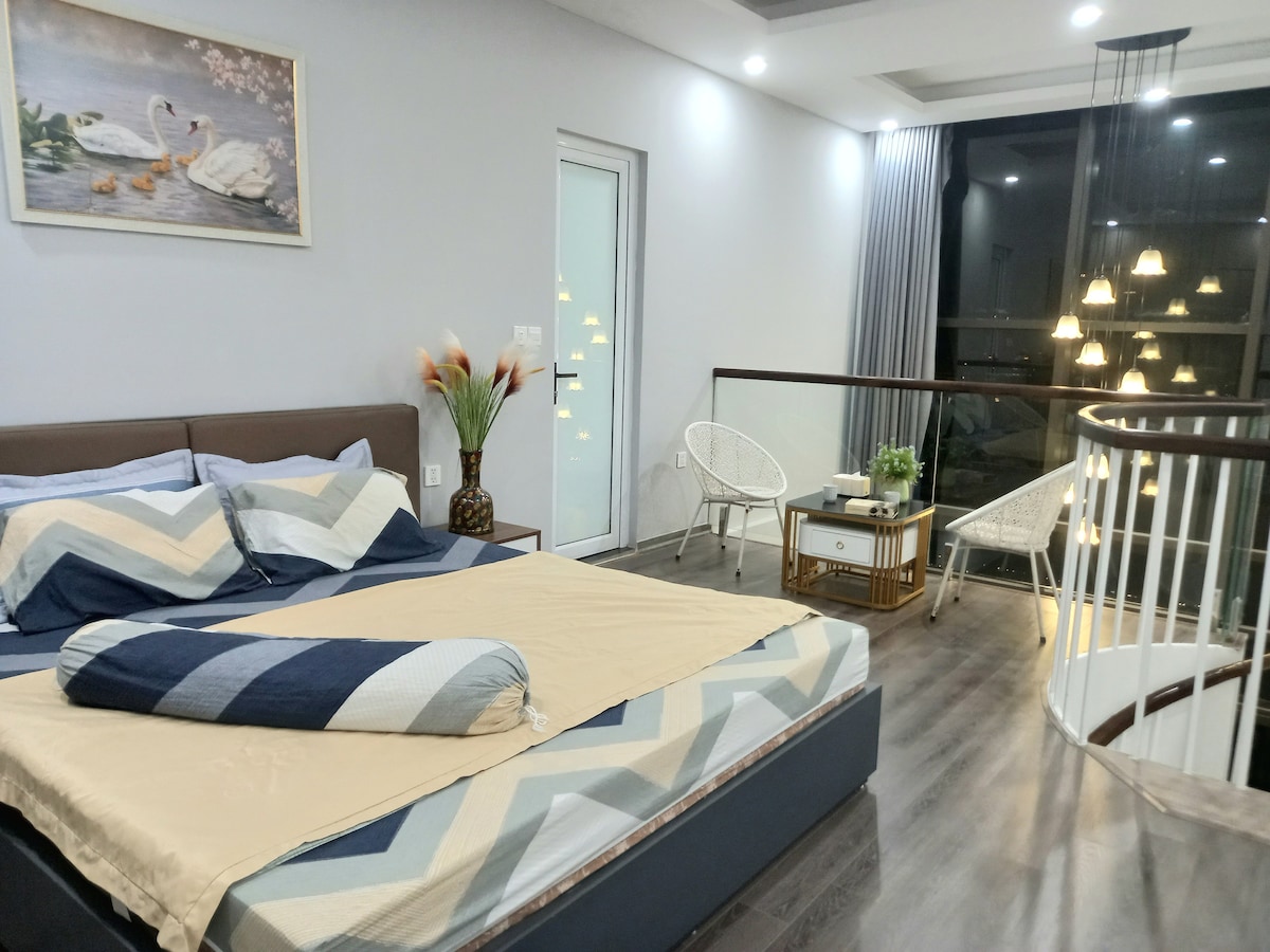 A warm studio bedroom is presented with a spacious bed featuring a light and dark blue patterned blanket. A large window offers a view of the outdoors, complemented by soft lighting from nearby fixtures. Seating includes stylish chairs beside a decorative table.