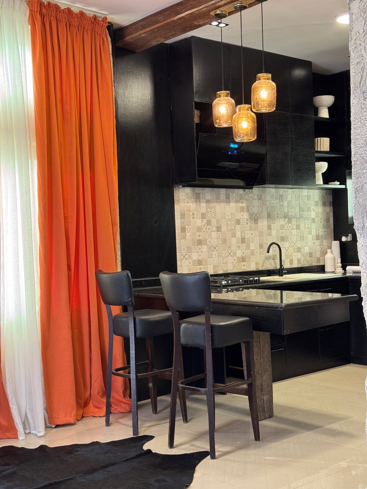 The kitchenette features dark cabinetry with modern fixtures and a textured backsplash. Two tall bar stools are positioned at the counter for seating. Warm, ambient lighting is provided by three pendant lamps, while bright orange curtains frame the window, adding a pop of color to the space.