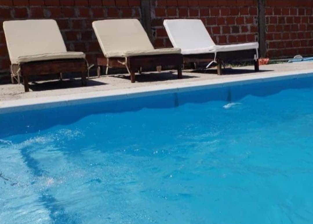 A clear swimming pool is featured, bordered by lounge chairs in light cream and white hues. The pool's water glistens under sunlight, offering a refreshing view. Bricks in a warm red tone create a simple backdrop behind the pool area.