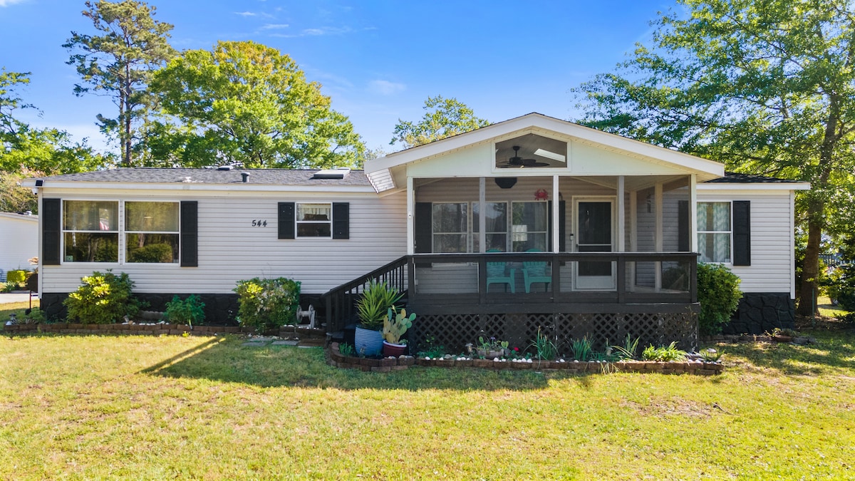 A well-maintained house features a welcoming porch with seating, surrounded by landscaped greenery. Large windows allow natural light to fill the space. The yard is neatly kept, with flower beds framing the area, providing an inviting view from the front.