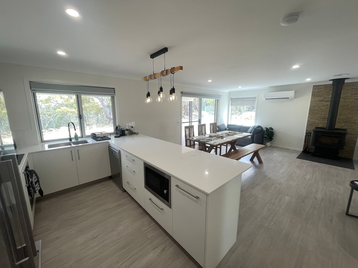 An open living area is highlighted by a modern kitchen featuring a large island with a built-in microwave. Natural light filters through multiple windows, illuminating the dining area and comfortable seating space. A wood heater is positioned in the corner, adding warmth to the environment.