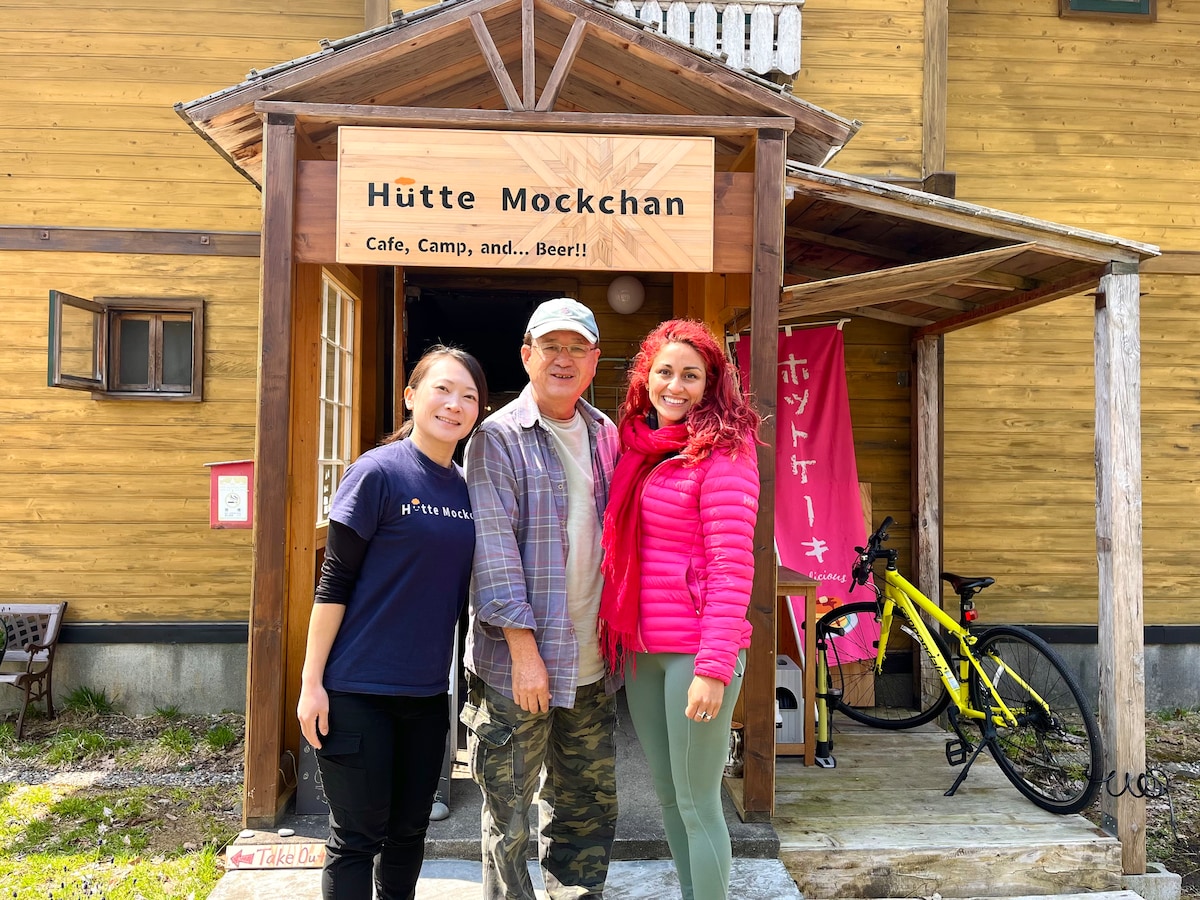 The entrance to Hütte Mockchan is showcased, featuring a wooden sign that reads 'Hütte Mockchan.' Two individuals stand alongside a third person, framed by a welcoming exterior. The rustic wooden structure is complemented by bicycles parked nearby and colorful banners gently fluttering in the breeze.