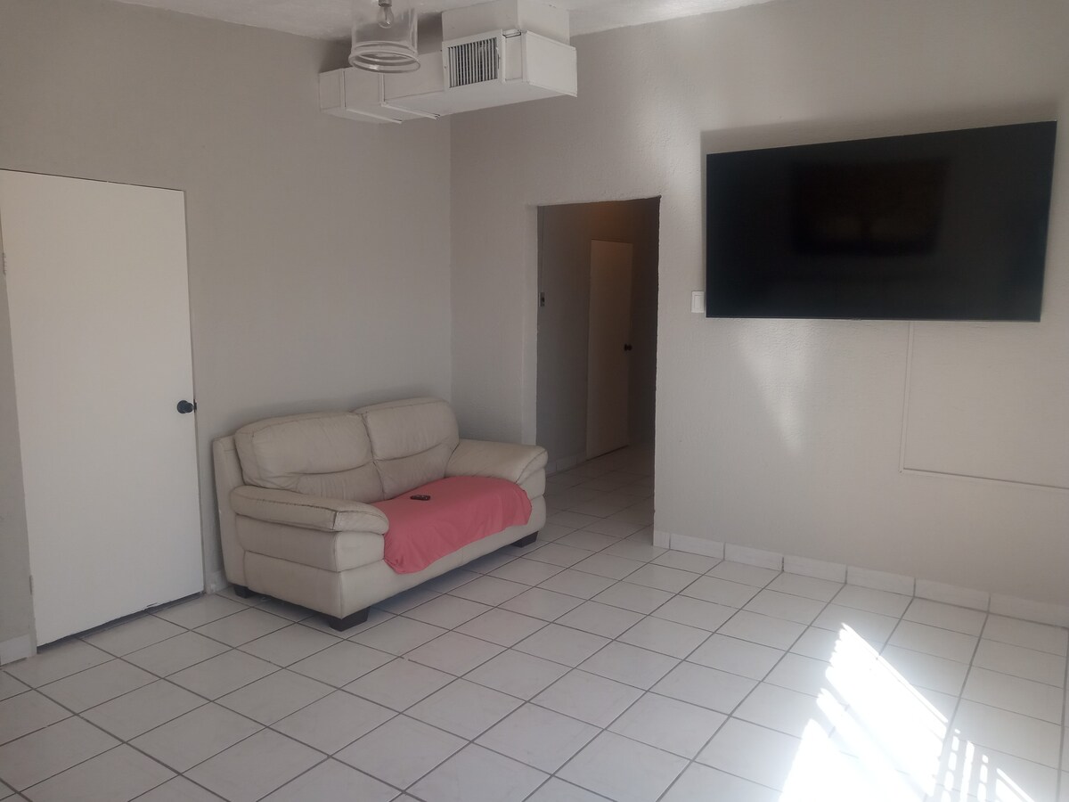 A light-filled living area features a cream-colored couch draped with a red throw, positioned against a neutral wall. A large flat-screen television is mounted above a simple console. Light tiles cover the floor, enhancing the spacious feel of the room.