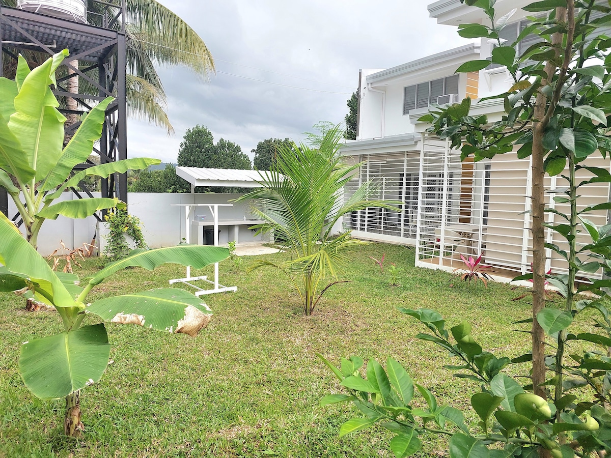 A well-maintained garden space is presented, featuring green grass and a variety of plants, including banana and palm trees. A white table sits under a shaded structure, providing a spot to enjoy the surroundings. The home is visible in the background, adding to the serene atmosphere.