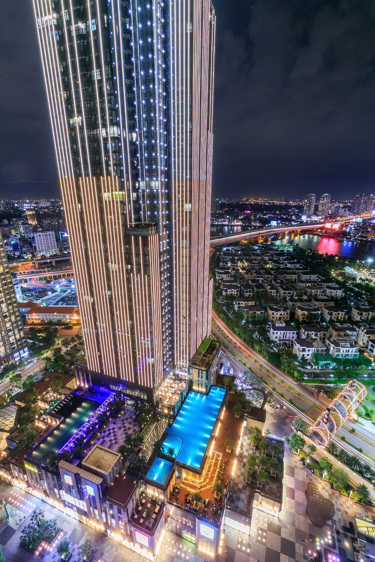 The image captures the Landmark 81 building at night, illuminated by vibrant lights. The view showcases the surrounding cityscape and waterways, with a large pool area visible on the building's lower levels, accentuating the modern architecture.