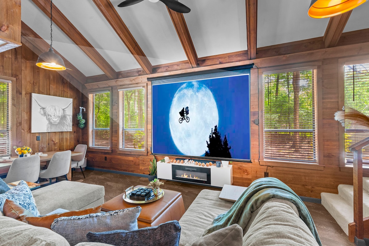 A cozy living area is centered around a comfortable sectional sofa, complementing a stylish coffee table. A large screen displays an artistic image against a natural backdrop, with large windows offering views of the surrounding woods. Wooden beams enhance the cabin's rustic charm.