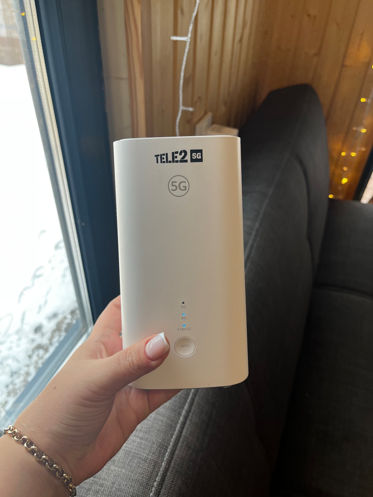 A white 5G router is held in a hand, displaying the brand name 'TELE2' along with a signal indicator. The background shows a dark grey sofa and a snowy outdoor scene visible through a nearby window.