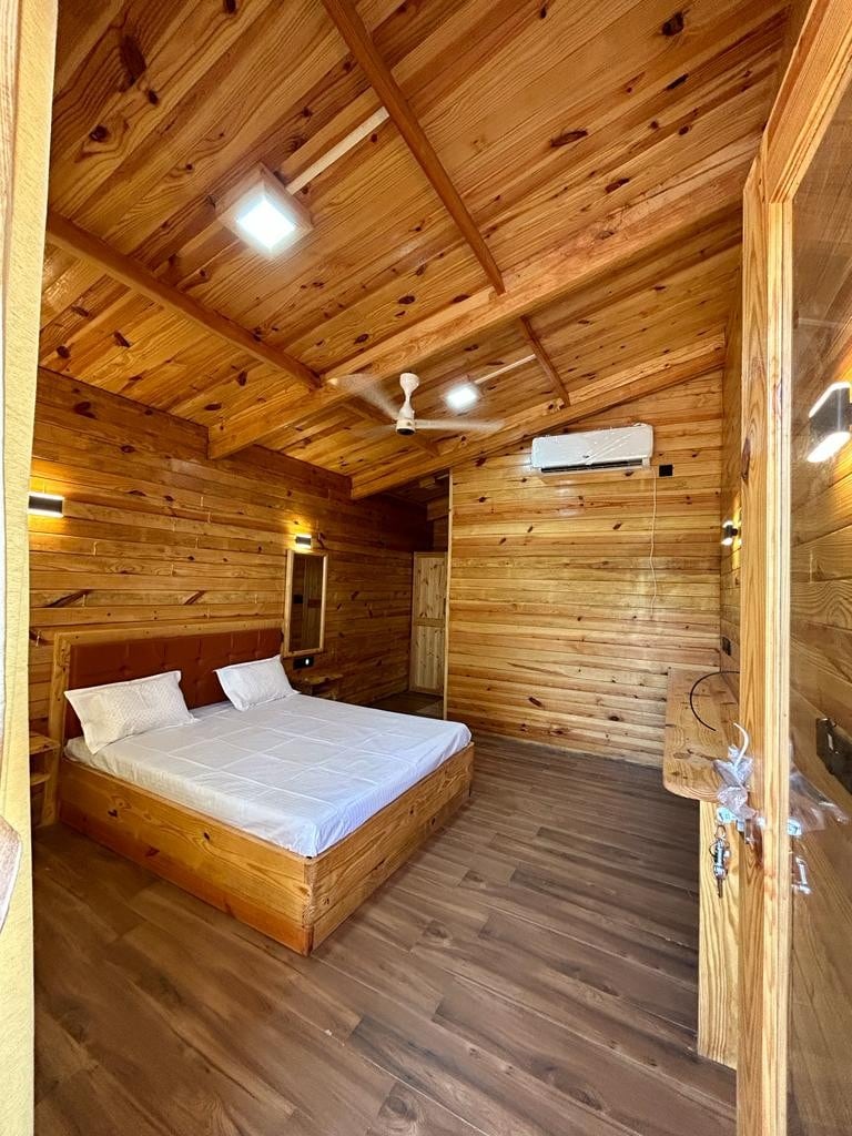 A room features wooden walls and ceiling, creating a natural aesthetic. A spacious bed with white linens is centered in the room. Soft lighting from wall fixtures contributes to a cozy ambiance. An air-conditioning unit is mounted above the bed for comfort.
