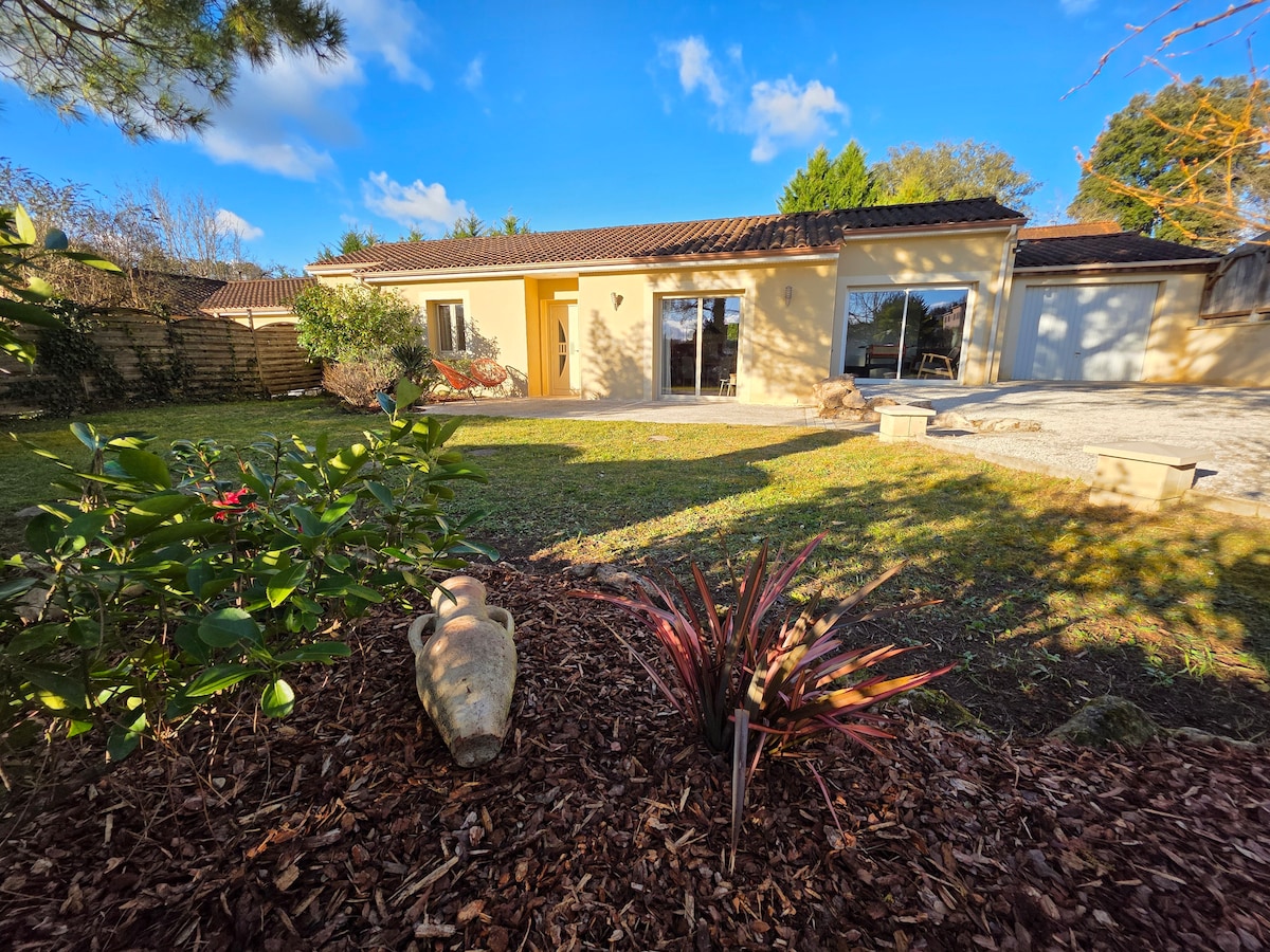 A single-story house is observed from the garden, featuring multiple large windows that reflect natural light. The garden includes manicured grass with green foliage, and a decorative pottery piece is complemented by vibrant plants, creating a serene outdoor space.