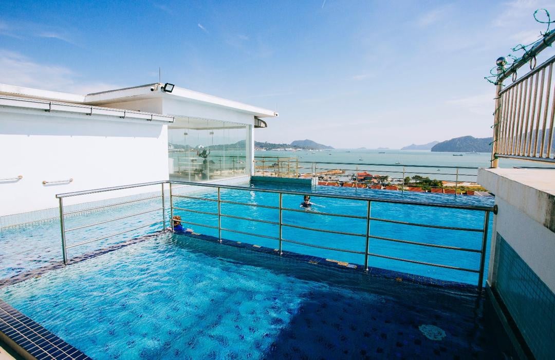 A rooftop pool is displayed with a clear view of the sea in the background. The water reflects sunlight, and a railing surrounds the pool area. Rolling hills and distant mountains create a scenic backdrop, enhancing the overall atmosphere of relaxation.