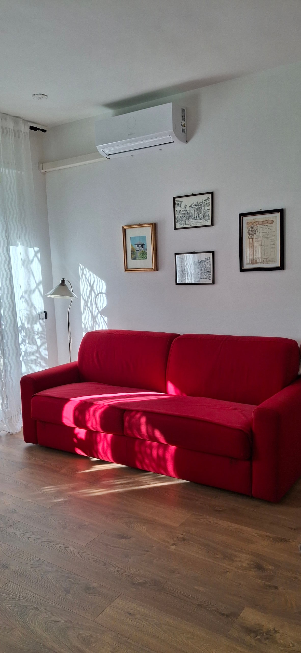 A bright living area features a red sofa that can be converted into a spacious bed. Natural light filters through sheer curtains, casting soft shadows on the wooden floor. Wall art and framed documents are displayed above the sofa, contributing to the room's character.