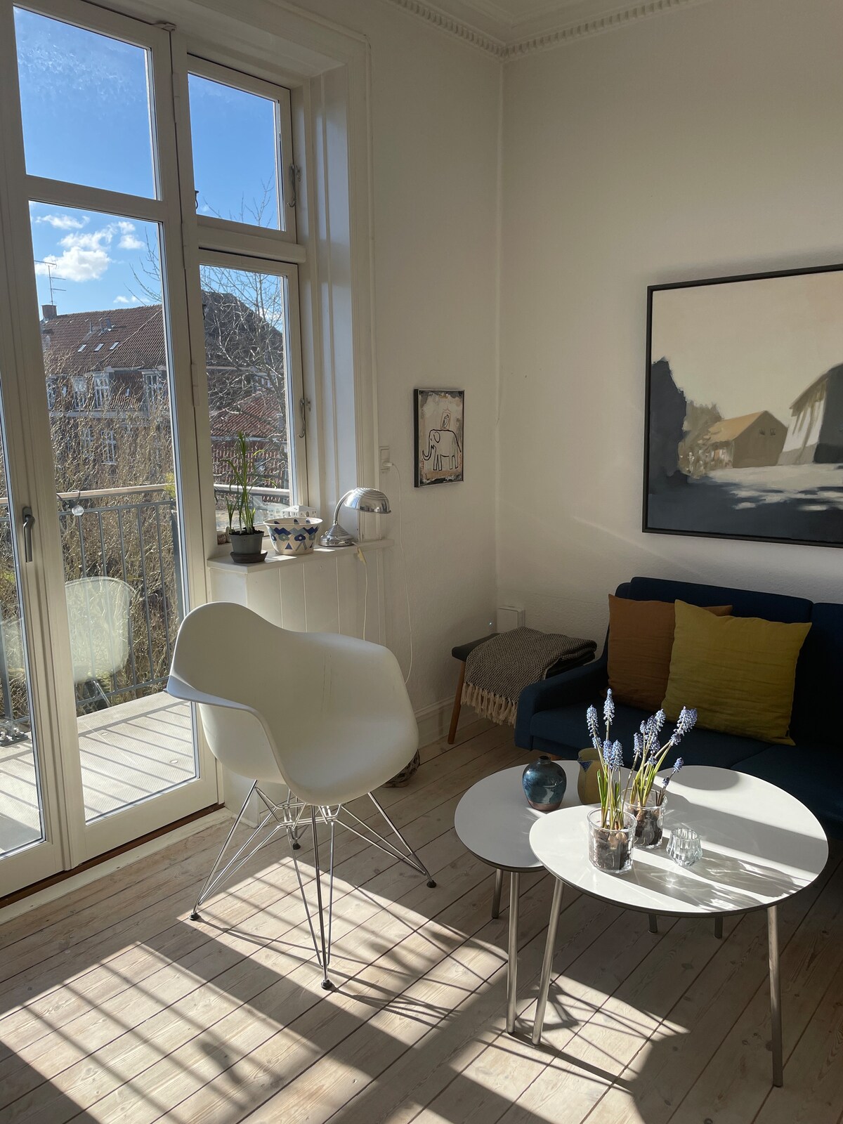 An airy living space is illuminated by natural light, featuring large windows that open to a small balcony. A white chair sits beside a minimalist table, adorned with decorative items. A dark blue sofa with colorful cushions adds comfort to the room, complemented by subtle artwork on the walls.