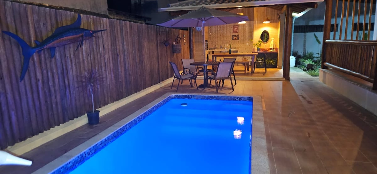 A tranquil pool area is highlighted with a vibrant blue water reflecting the surrounding lights. Lounge chairs are placed near the pool. In the background, an outdoor dining area is visible under a shaded pergola, with a barbecue and a decorative fish mounted on the wall.