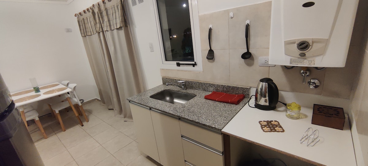A fully equipped kitchen is presented featuring a granite countertop, a stainless steel sink, and essential appliances, including a kettle and a microwave. Cooking utensils are arranged on the wall. A dining table with chairs is visible in the background, creating a functional and inviting space.