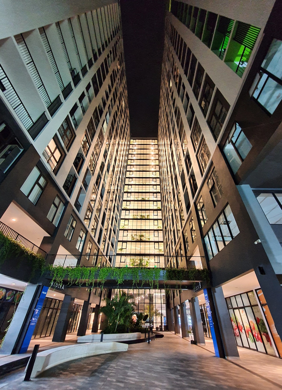 The image captures a modern building complex illuminated at night, showcasing tall, sleek facades with numerous windows. Greenery decorates the entrance area, and the courtyard is highlighted by ambient lighting, creating a vibrant atmosphere in the urban setting.