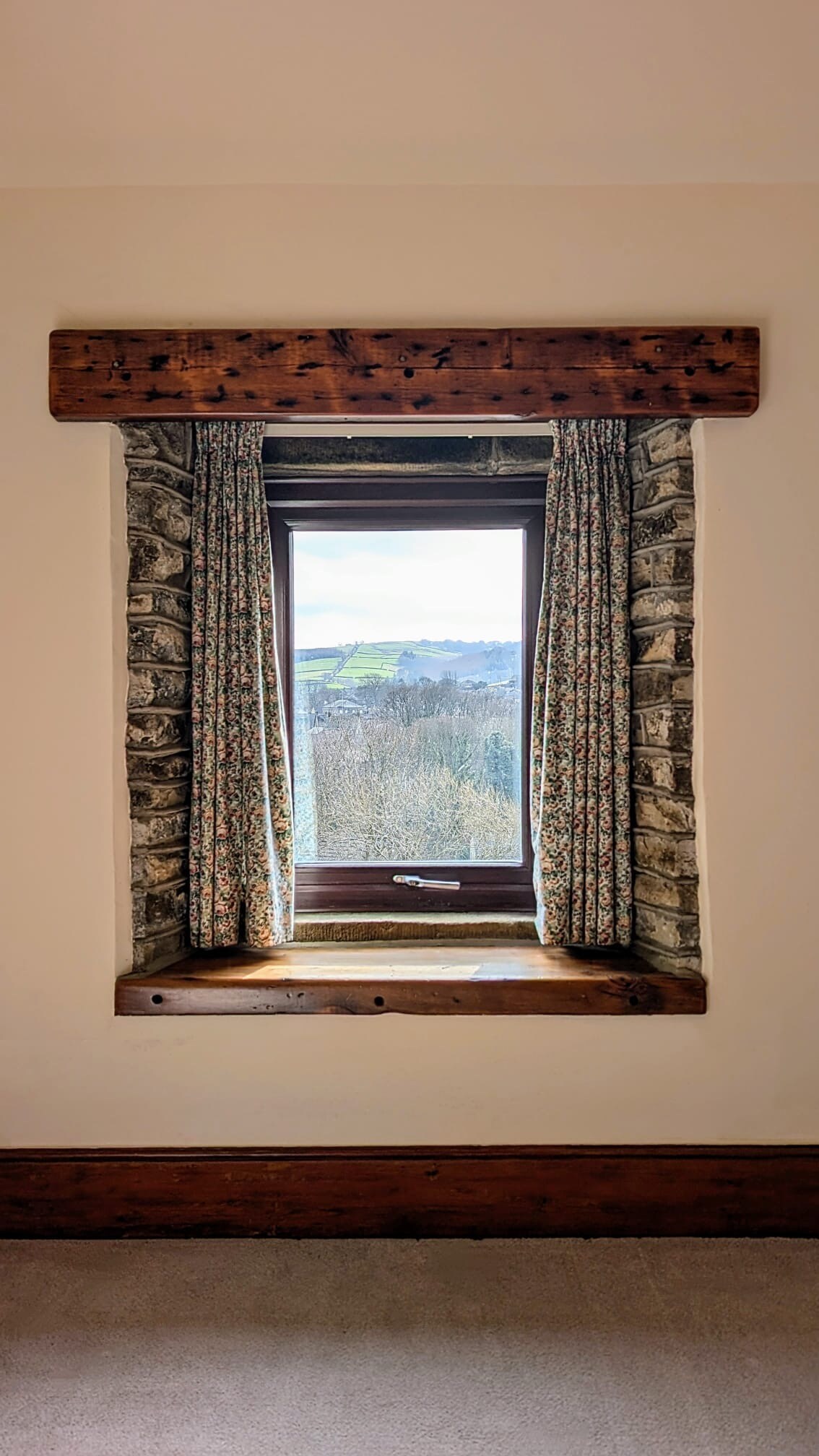 A window framed by rustic wooden trim is highlighted, showcasing views of the surrounding countryside. Floral curtains adorn the window, complementing the natural light that filters through. The stone wall adds a touch of historic character to the space.