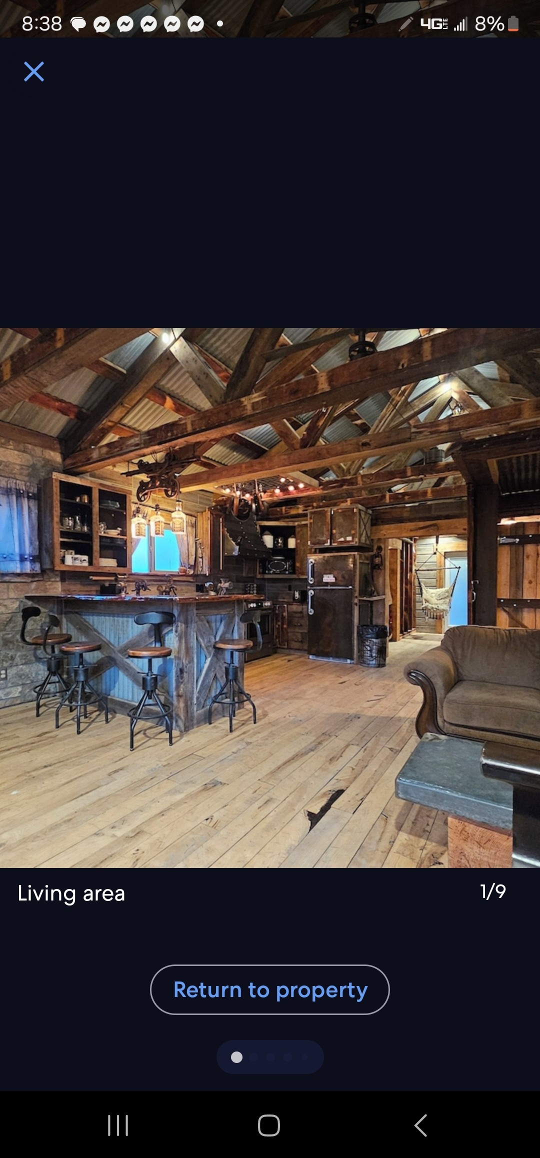 The living area features a rustic wooden bar with high stools, surrounded by exposed wooden beams and a stone wall. A cozy couch is positioned nearby, while warm light fixtures create a welcoming environment. A glimpse of the kitchen area is visible in the background.