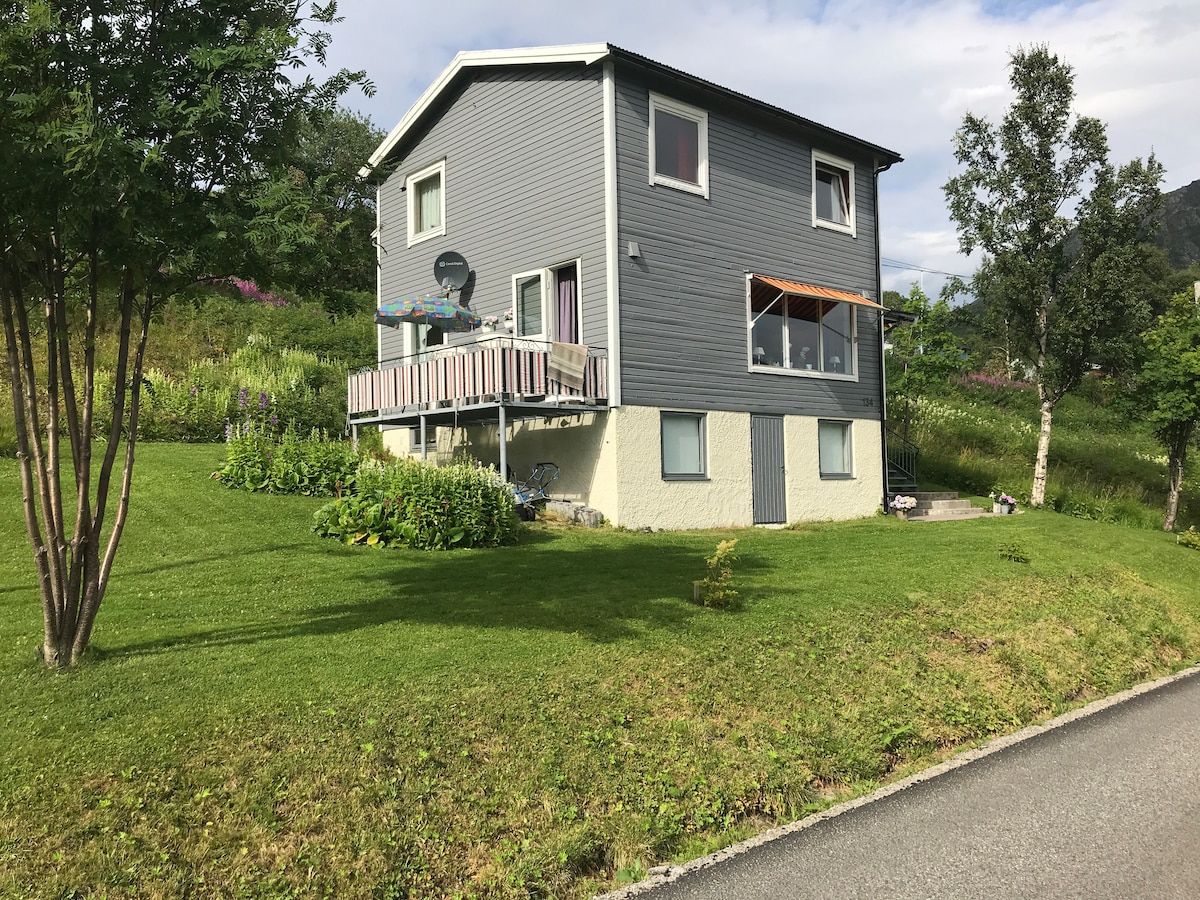 A two-story grey house is situated on a green lawn, surrounded by trees and shrubs. Large windows offer views of the landscape, while a balcony is equipped with a clothesline and outdoor seating. The setting reflects a peaceful, natural environment.