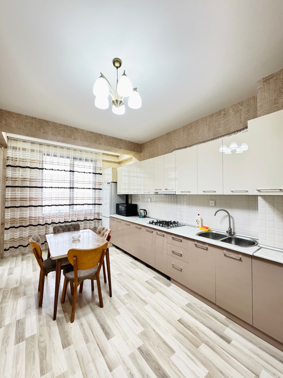 A bright kitchen area is featured, equipped with modern appliances and ample cabinetry. A dining table with four chairs is set, with light streaming in through large windows adorned with sheer curtains. The soft-toned walls enhance the clean and functional space.