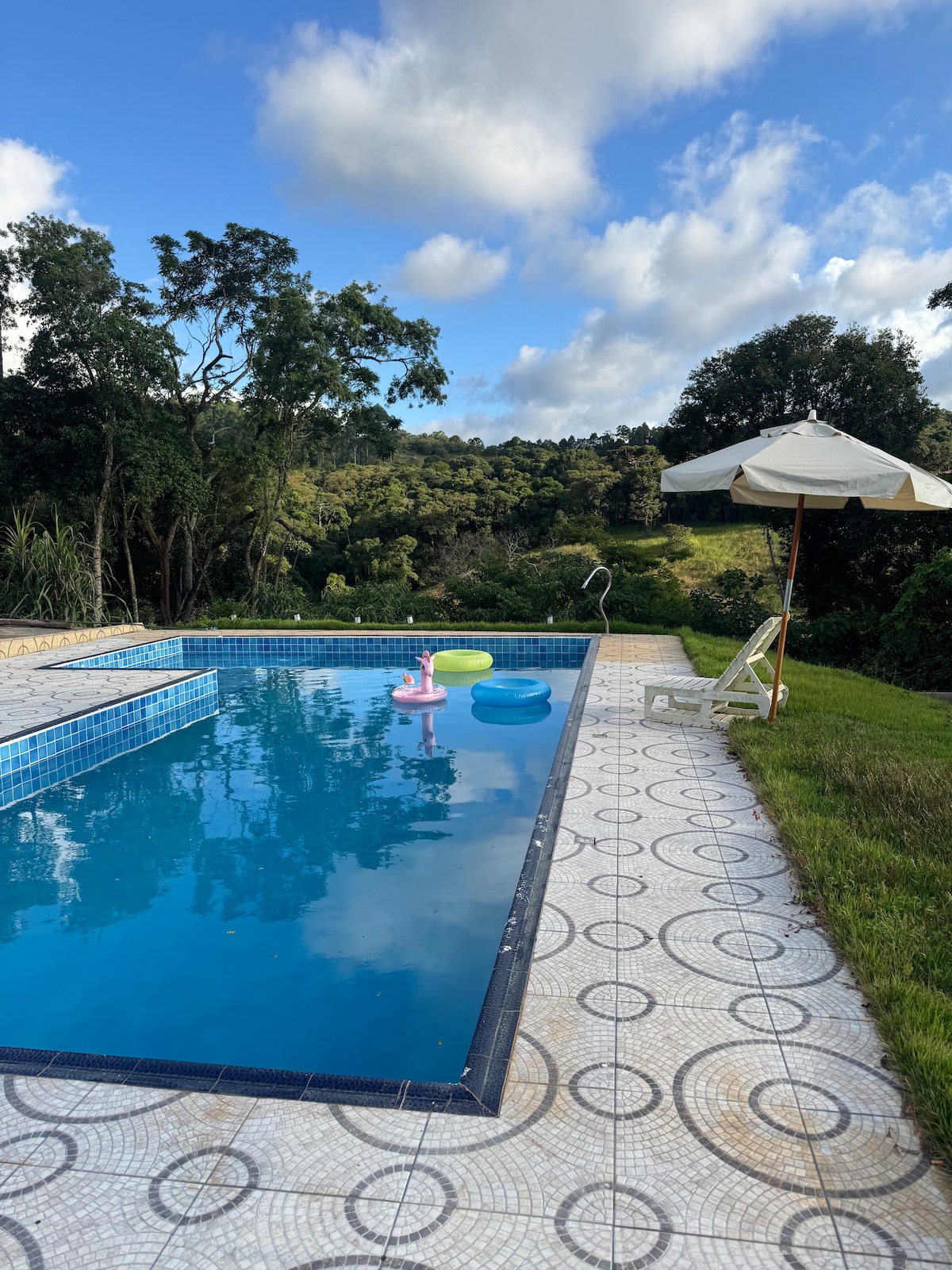 A refreshing swimming pool is surrounded by lush greenery, inviting relaxation. Two inflatable floats are visible in the water, with a shaded area provided by a large umbrella and a lounge chair nearby. Scenic hills can be seen in the background under a partly cloudy sky.