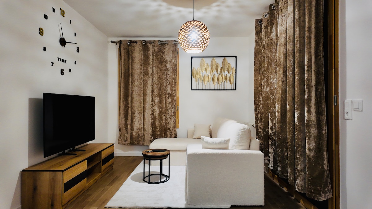 A cozy living area is featured with a light-colored sofa and a round coffee table. A television is positioned on a wooden TV stand. Soft curtains frame the window, and a decorative wall clock adds a unique touch to the space.