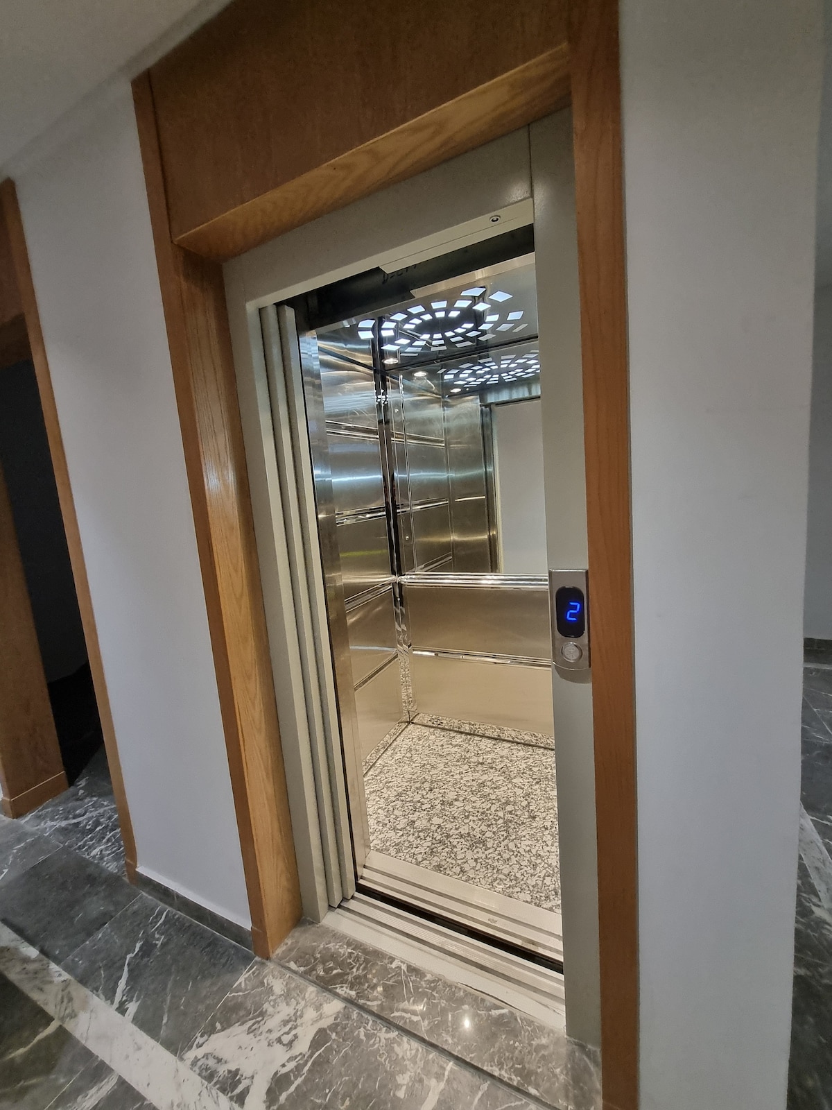 An elevator is shown within a modern building entrance. The interior features a sleek silver finish, mirrored walls, and a decorative ceiling pattern. A digital display indicates the current floor, and the surrounding area is enhanced by polished marble flooring.