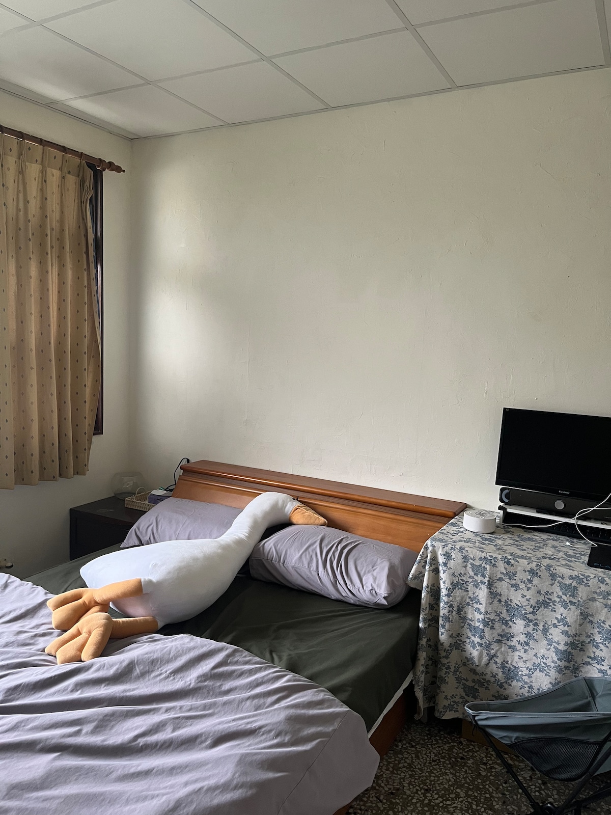 A simple sleeping area features a wooden headboard with two pillows on a bed. A plush goose toy rests comfortably on the gray bedding. A small desk with a monitor is situated beside the bed, and a patterned fabric drapes over the desk.