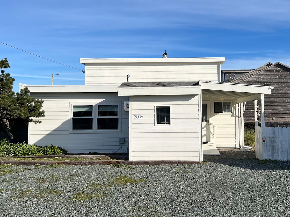 A light-colored beach bungalow is presented, featuring a covered entrance and a gravel driveway. Large windows allow natural light to enter the interior, while surrounding greenery adds a touch of nature to the charming façade.