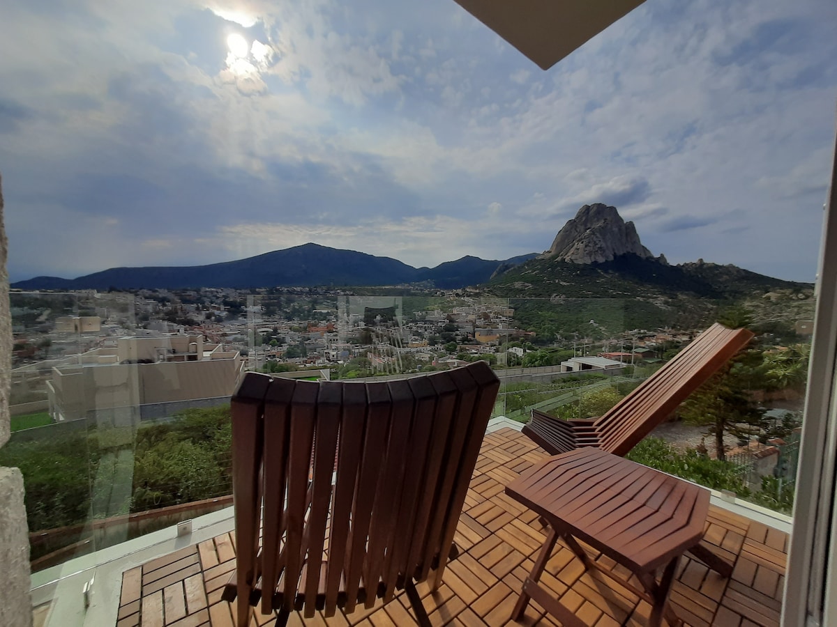 A spacious outdoor terrace features wooden lounge chairs overlooking a panoramic view of the Peña de Bernal and surrounding mountains. A clear sky with wispy clouds adds to the serene atmosphere, while warm sunlight casts gentle shadows on the tiled floor.