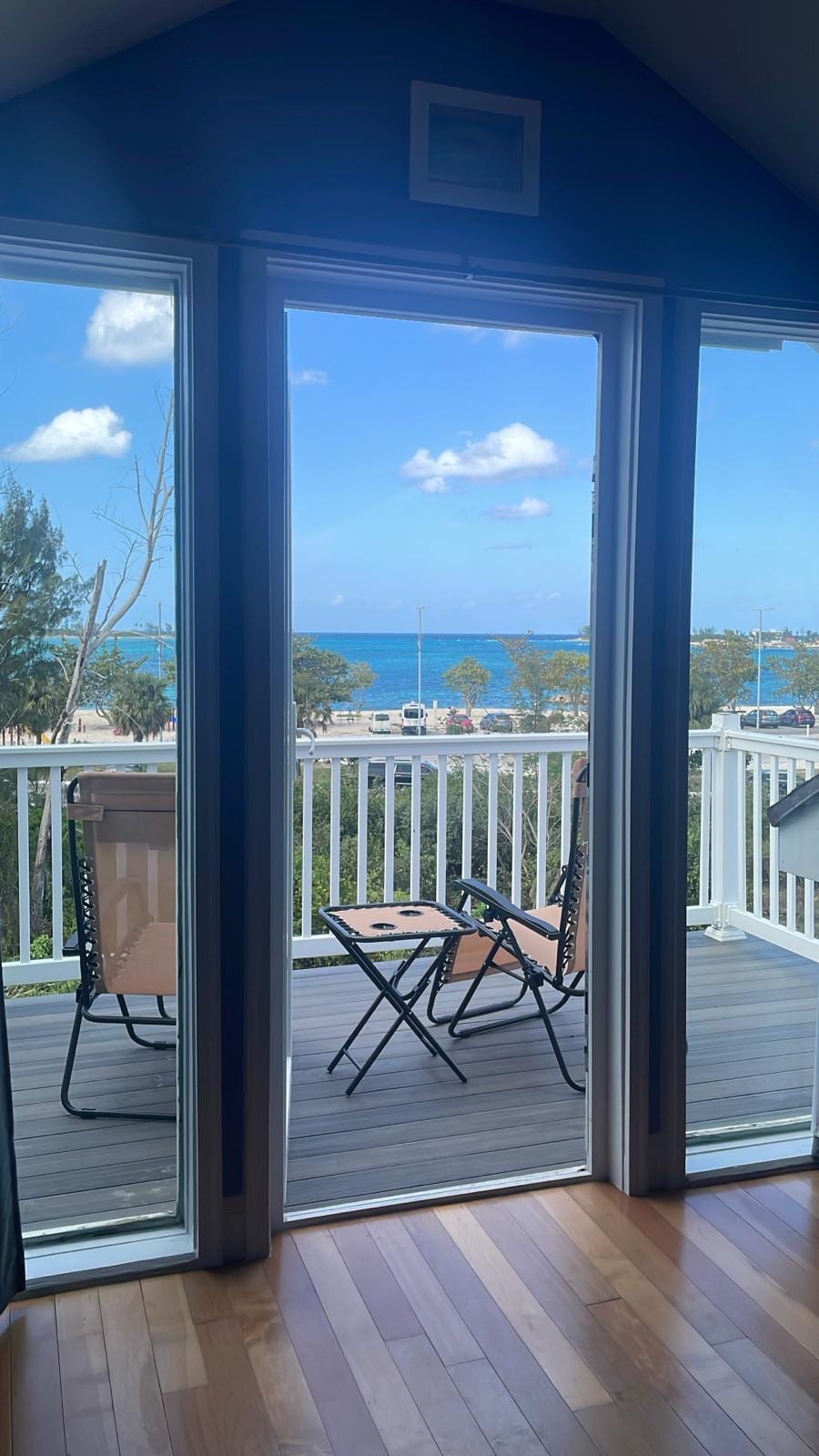 A balcony area is presented through expansive glass doors, featuring two chairs and a table. The scene is framed by lush greenery and showcases a panoramic view of the sparkling sea and distant shoreline under a blue sky with scattered clouds.